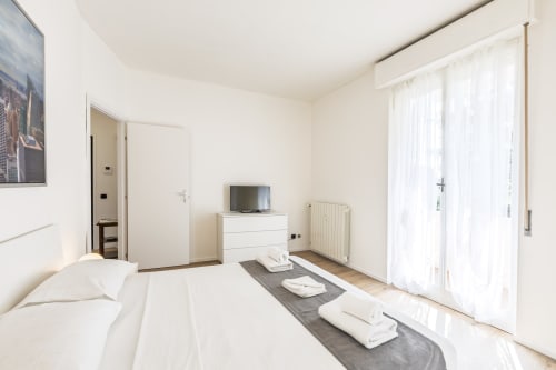 Solumnia Urban Nest, bilocale a Milano con letto matrimoniale, comò bianco con TV e porte in vetro verso il balcone con vista città