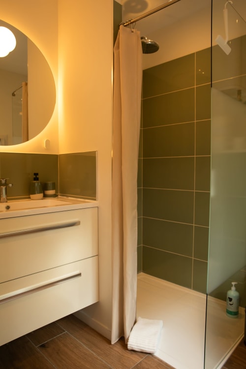 Salle de bain moderne de la chambre d'hôtes Recevoir par Charline B&B à Biganos avec vasque blanche, miroir rond lumineux, carrelage vert olive et douche à l'italienne