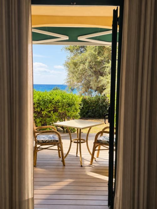 Terrasse de l'Hôtel Thalassa à Cargèse avec table de bistro, chaises et vue sur la Méditerranée au-delà de la végétation côtière
