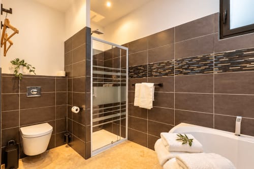 Salle de bain moderne de l'hôtel La Villa Monarque à Cap d'Agde avec carrelage brun, douche vitrée, baignoire et sèche-serviettes électrique