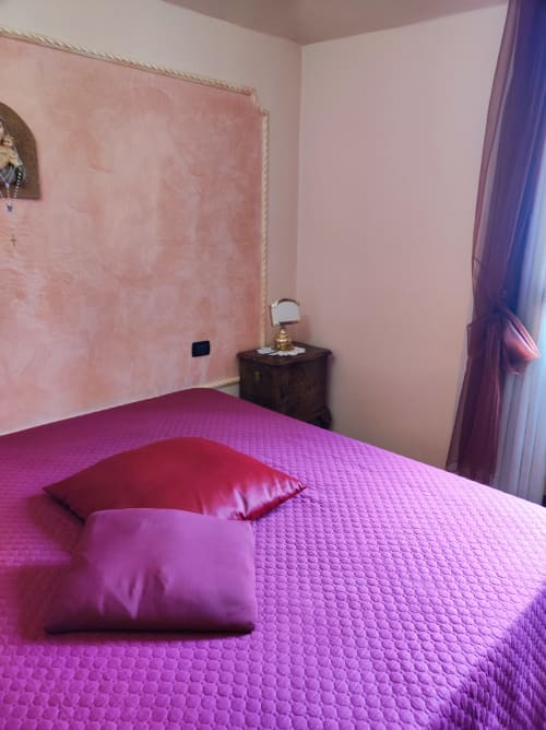 Camera da letto in rosa brillante presso Gruppo Platone Alessandria con biancheria magenta, cuscini, comodino in legno scuro e tende viola