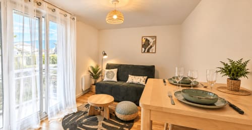 Studio moderne chez NN Patrimoine Douvaine avec table à manger, canapé anthracite, suspension en rotin et balcon vue sur la ville