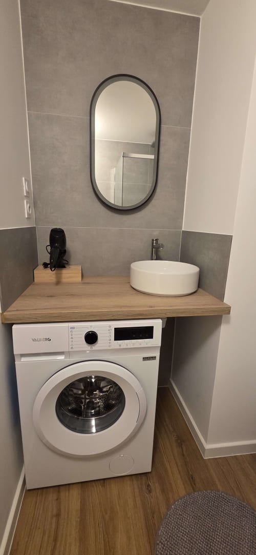 Buanderie moderne avec lave-linge, plan de travail en bois, lavabo blanc et miroir ovale à l'hôtel NN Patrimoine de Douvaine