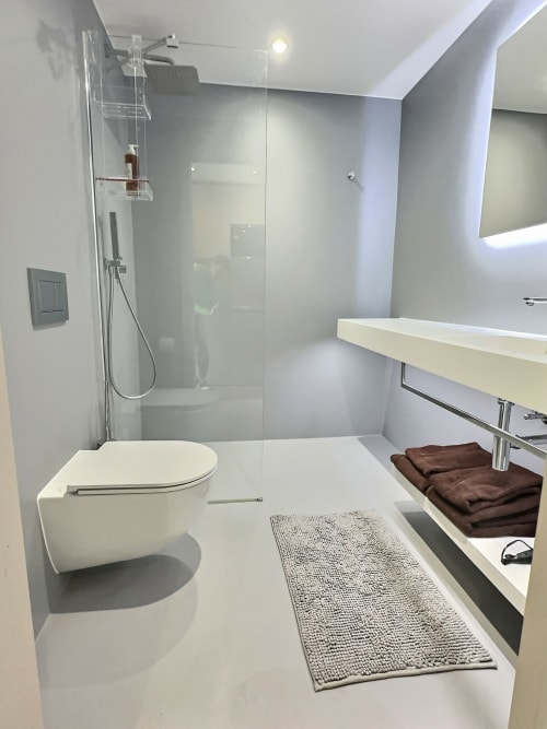 Modern studio bathroom at Attica Résidences with white vanity, wall-mounted toilet, rainfall showerhead, and gray bath mat.