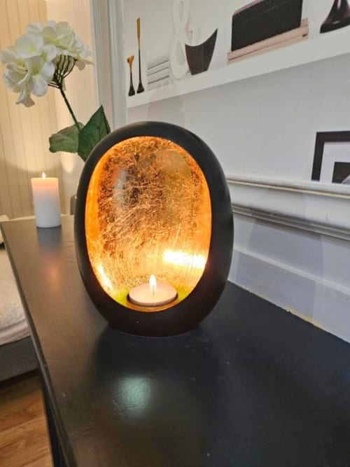 Lampe ovale noire moderne avec intérieur orange luminescent et bougie chauffe-plat, à côté d'une bougie blanche et d'une fleur blanche dans la décoration d'un appartement