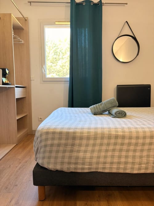Chambre moderne au Clos d'Aix avec lit queen-size, armoire bois, miroir rond et vue jardin par rideaux turquoise