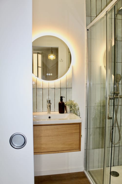 Salle de bain moderne de la suite Mi Casa Es Su Casa à Carcassonne, avec miroir LED circulaire, meuble vasque en bois et douche vitrée.