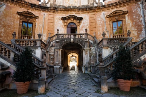 Sicilian palazzo courtyard at Sikania Agri Relais in Caccamo with stone staircases, ornate balconies, and terracotta planters