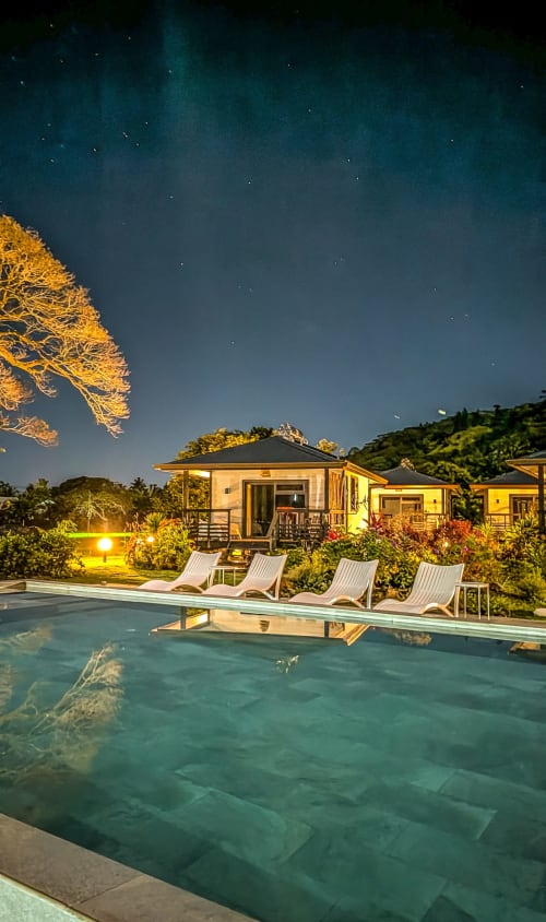 Sublime Tahiti Beach Retreat bungalows at night with illuminated pool, loungers, and starry sky over tropical landscape