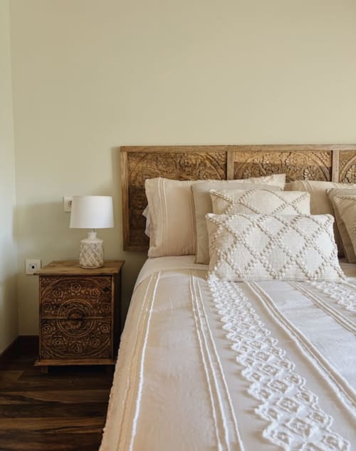 Dormitorio rústico en Alojamientos Montaña Leonesa con cabecero de madera tallada, ropa de cama color crema y mesilla ornamentada con lámpara.