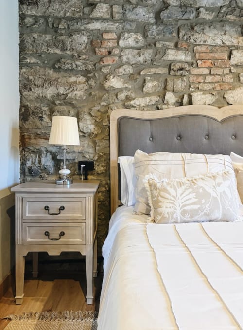 Acogedora habitación en Alojamientos Montaña Leonesa con pared de piedra vista, cama tapizada en crema y mesita de noche con lámpara
