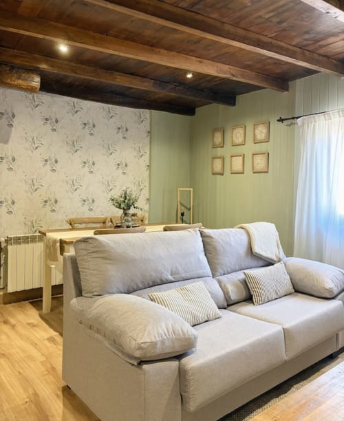 Acogedor dormitorio con sofá crema, vigas de madera vista, papel pintado floral, paredes verdes y vistas al jardín en Alojamientos Montaña