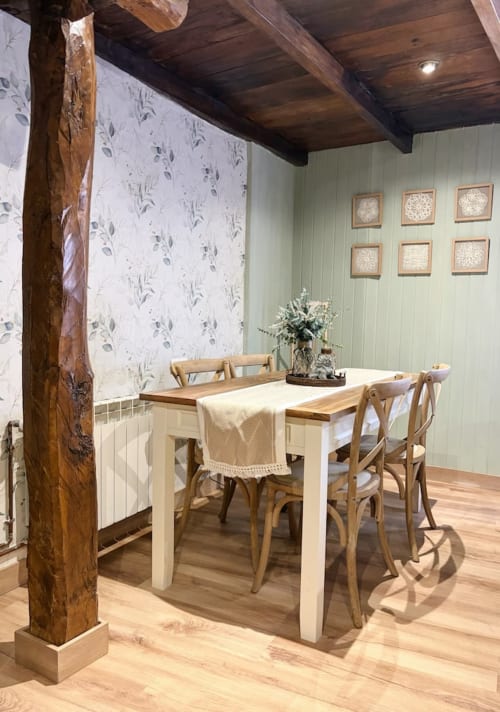 Comedor rústico en Alojamientos Montaña Leonesa con vigas de madera, mesa blanca, sillas naturales y papel tapiz con vistas al jardín