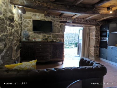 Sala de estar con muros de piedra, sofá de cuero negro, televisor, vigas de madera y vistas al jardín en Bikarte Ribeira Sacra, Sober