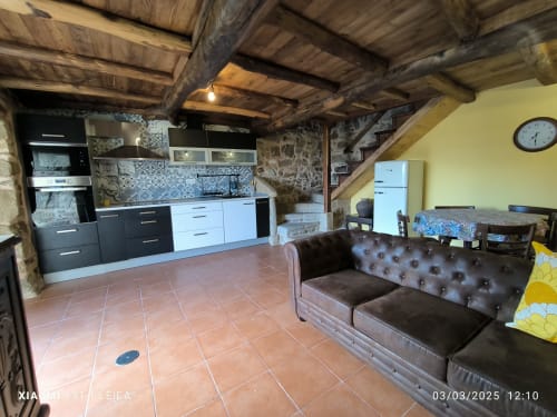 Área de estar diáfana y rústica en Bikarte Ribeira Sacra con cocina moderna, vigas de madera, baldosas de terracota y cuero marrón
