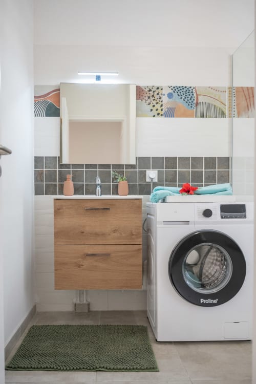 Modern laundry bathroom with wood vanity, washing machine, gray tile backsplash, and decorative accessories at Vista Del Mar