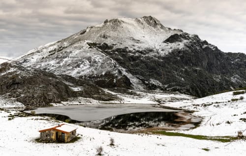 Paisaje montañoso cubierto de nieve con lago congelado y cabaña amarilla en Alojamientos Montaña Leonesa, Cofiñal