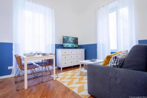 Camera moderna con pareti azzurre, pavimento in legno, scrivania, cassettiera con TV e divano grigio a Seven Suites Sanremo