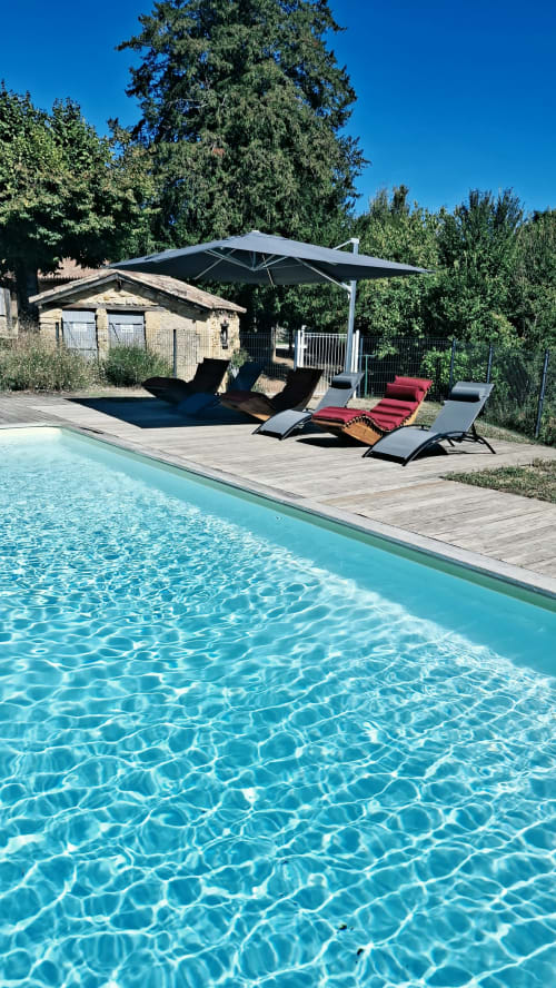 Piscine turquoise aux Gîtes Le Pech avec chaises longues, parasol débordant et maison en pierre à Sainte-Foy-de-Longas
