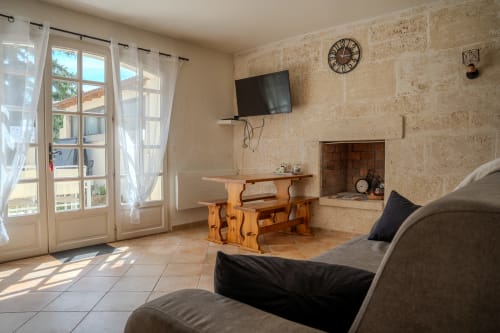 Séjour lumineux du gîte Au Coin du Feu avec murs en pierre, table en bois, ancienne cheminée et vue sur le jardin