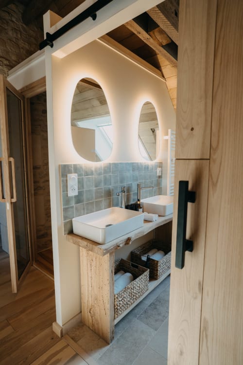 Modern bathroom vanity with dual vessel sinks, illuminated oval mirrors, and woven storage baskets at Le Clos d'Omaha hotel