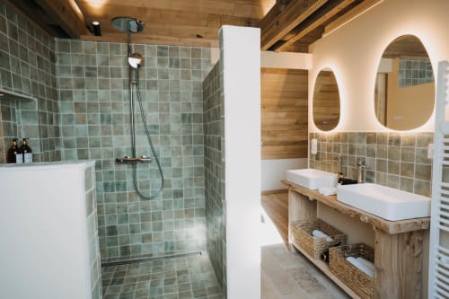Modern bathroom at Le Clos d'Omaha with green tile shower, dual white sinks, illuminated mirrors, wooden accents, and woven