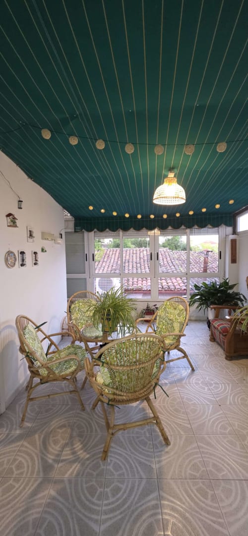 Terraza de apartamento iluminada por el sol con sillas de mimbre, techo verde, lámparas colgantes y vistas de tejados en Monforte de Lemos