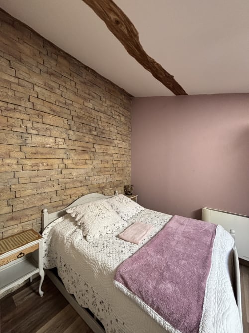 Chambre cosy du Mas Provençal avec mur en briques apparentes, lit blanc, couverture violette et plafond aux poutres en bois à Bollène