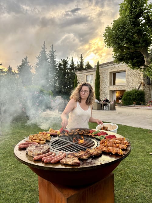 Femme grillant de la viande à La Petite Plaine B&B à Clansayes, avec villa en pierre et conifères au coucher de soleil