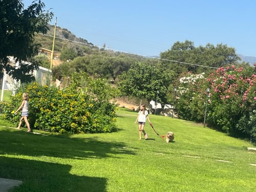 Bambini e cane giocano nel rigoglioso giardino di Resort Baia Cea, Bari Sardo, circondati da fiori e viste collinari