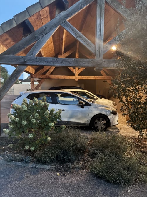Voiture blanche garée sous un carport en bois orné de plantes au gîte Le Clos d'Émilie, Saint-Marcel