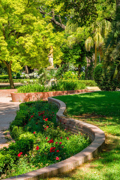 Exuberante jardín del Hostal San Vicente II en Sevilla con flores rojas, setos bien cuidados, palmeras y estatua