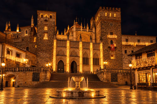 Plaza medieval nocturna con iglesia gótica iluminada, edificios de piedra y fuente en Guadalupe, Cáceres