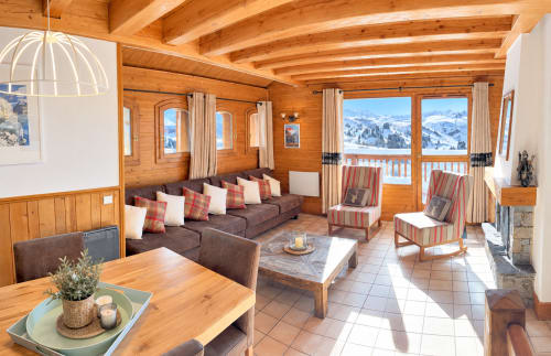 Salon cosy d'un refuge alpin avec poutres en bois, vue sur les montagnes, canapé marron, cheminée et balcon