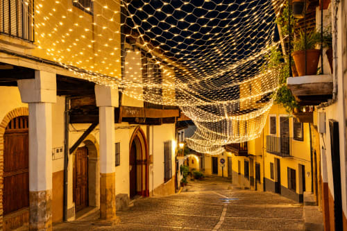 Encantadora calle empedrada en Guadalupe, Cáceres iluminada con mallas de luces doradas, mostrando la arquitectura tradicional española