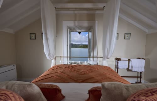 Quarto luminoso com vista para o lago na Casa da Lagoa, Furnas. Cama com roupa de cama terracota de frente para portas abertas com vista panorâmica