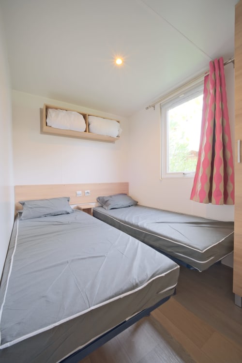 Chambre lumineuse avec trois lits, têtes de lit en bois, rideaux à motifs roses et fenêtre à Annick Mobilhome, Vias Plage