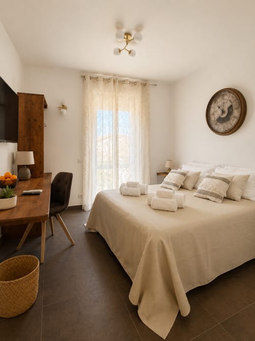 Camera spaziosa all'hotel Habemus di Rocca di Papa con biancheria color crema, scrivania in legno e vista su monumenti attraverso tende leggere