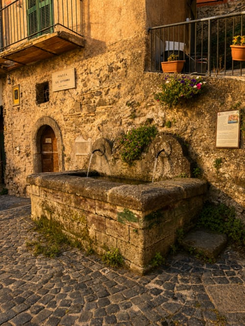 Pozzo in pietra storico con due archi in Piazza Vecchia, Rocca di Papa, circondato da edifici antichi e fiori colorati