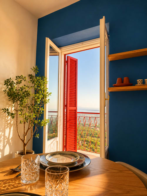 Luminosa sala da pranzo dell'hotel Habemus con pareti blu, tavolo in legno, vista lago dalle persiane rosse aperte e una pianta in vaso