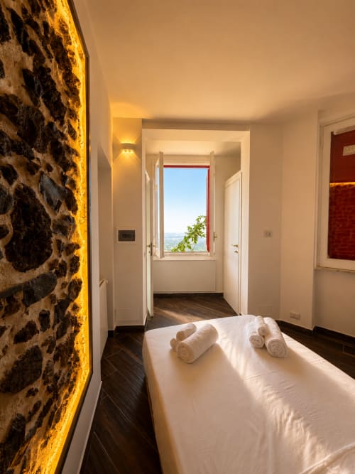 Camera luminosa dell'Habemus Rocca di Papa con parete in pietra, letto bianco, asciugamani arrotolati e vista lago dalla finestra
