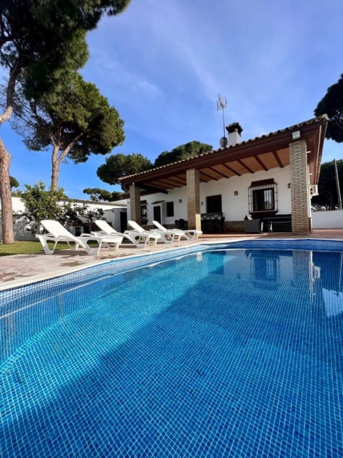 Piscina de la villa Barrosa con tumbonas, villa blanca y árboles mediterráneos bajo cielo azul