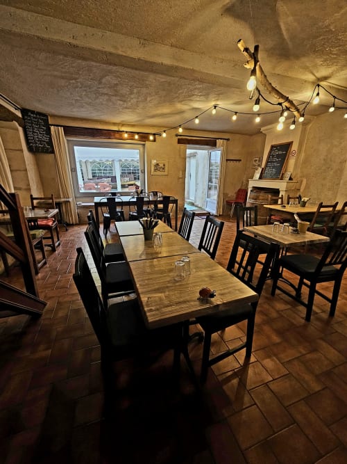 Salle à manger rustique du Logis de Chenac avec tables en bois, chaises noires, guirlandes lumineuses et vue sur la campagne