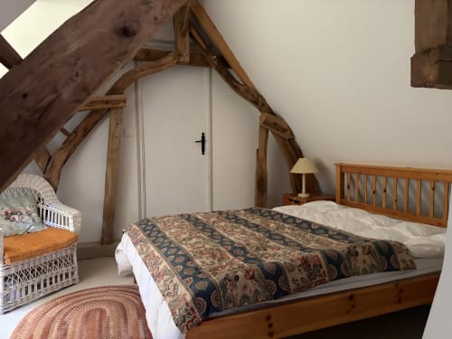 Chambre rustique mansardée avec poutres en bois apparentes, lit en bois, courtepointe à motifs et fauteuil en rotin