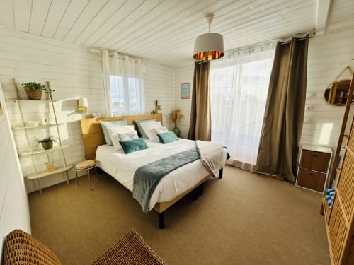 Chambre lumineuse à La Villa Plein Vent avec murs en bois blanc, lit queen, oreillers bleu canard, lampe suspendue et jardinage