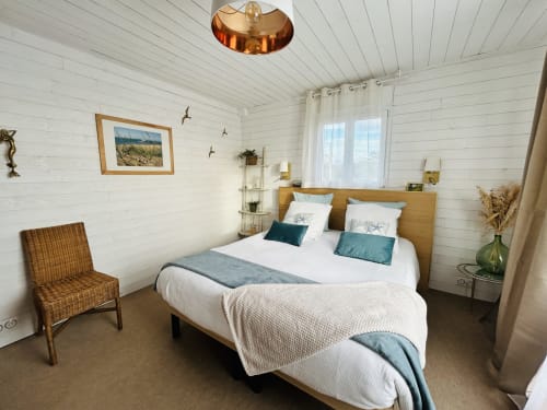 Chambre cosy à La Villa Plein Vent avec lit en bois, fauteuil en osier, murs blanc crème et vue sur le jardin