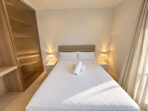 Dormitorio luminoso en apartamento Holibai de Baiona con cama queen, ropa blanca, iluminación cálida y mobiliario moderno