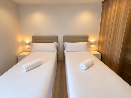 Camas gemelas con sábanas blancas en luminoso apartamento Holibai, Baiona. Incluye lámparas de mesita, cabeceros y cortinas de madera.