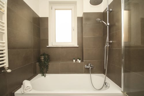 Modern bathroom in Palermo apartment with white bathtub, brown tile walls, rainfall showerhead, and natural window light