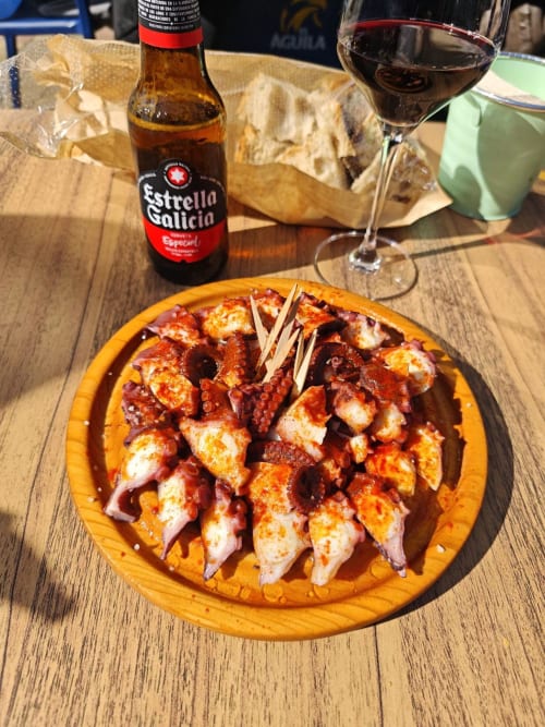 Brochetas de pulpo a la parrilla en plato amarillo con cerveza Estrella Galicia y vino tinto en Holibai, Baiona